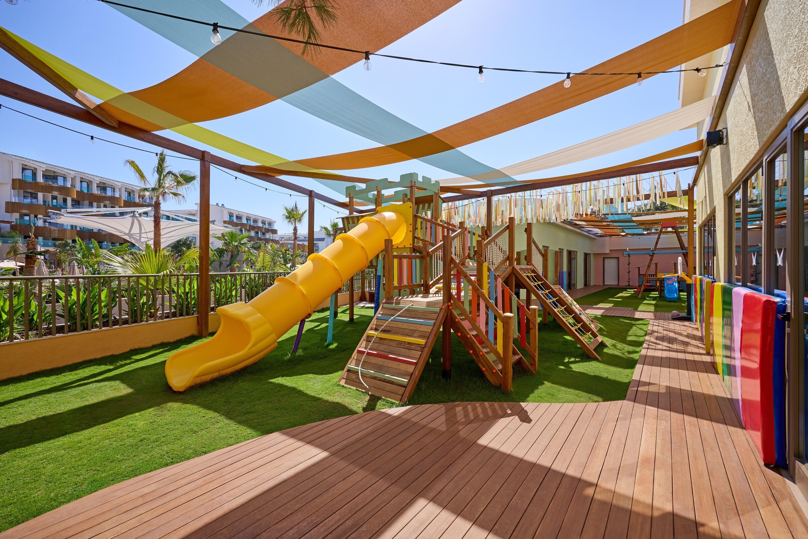 A children's play area with a yellow slide, wooden climbing structures, and artificial grass.