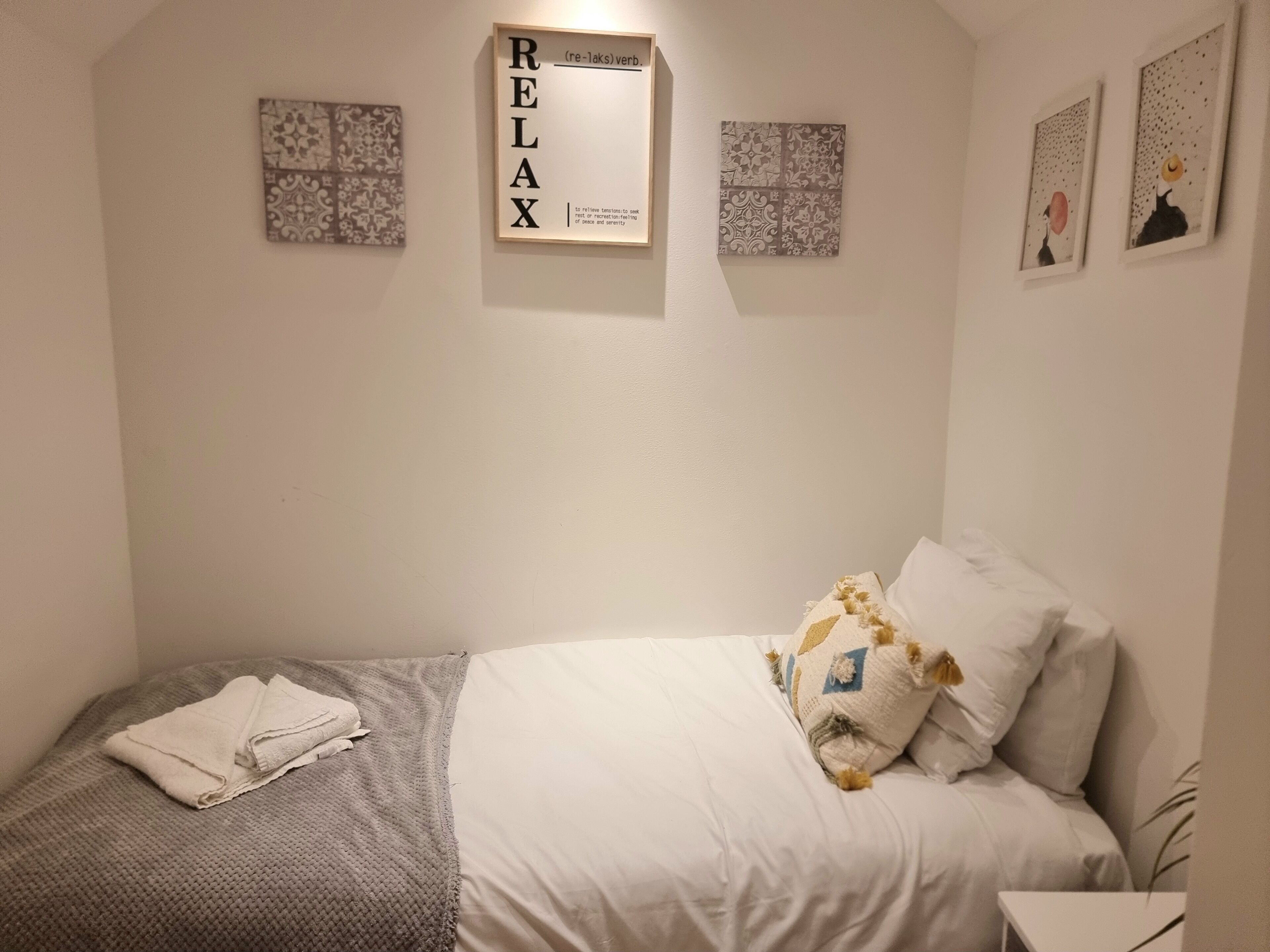A neatly made bed with a white comforter and pillows, a folded towel on the side, and a framed 'RELAX' sign on the wall.