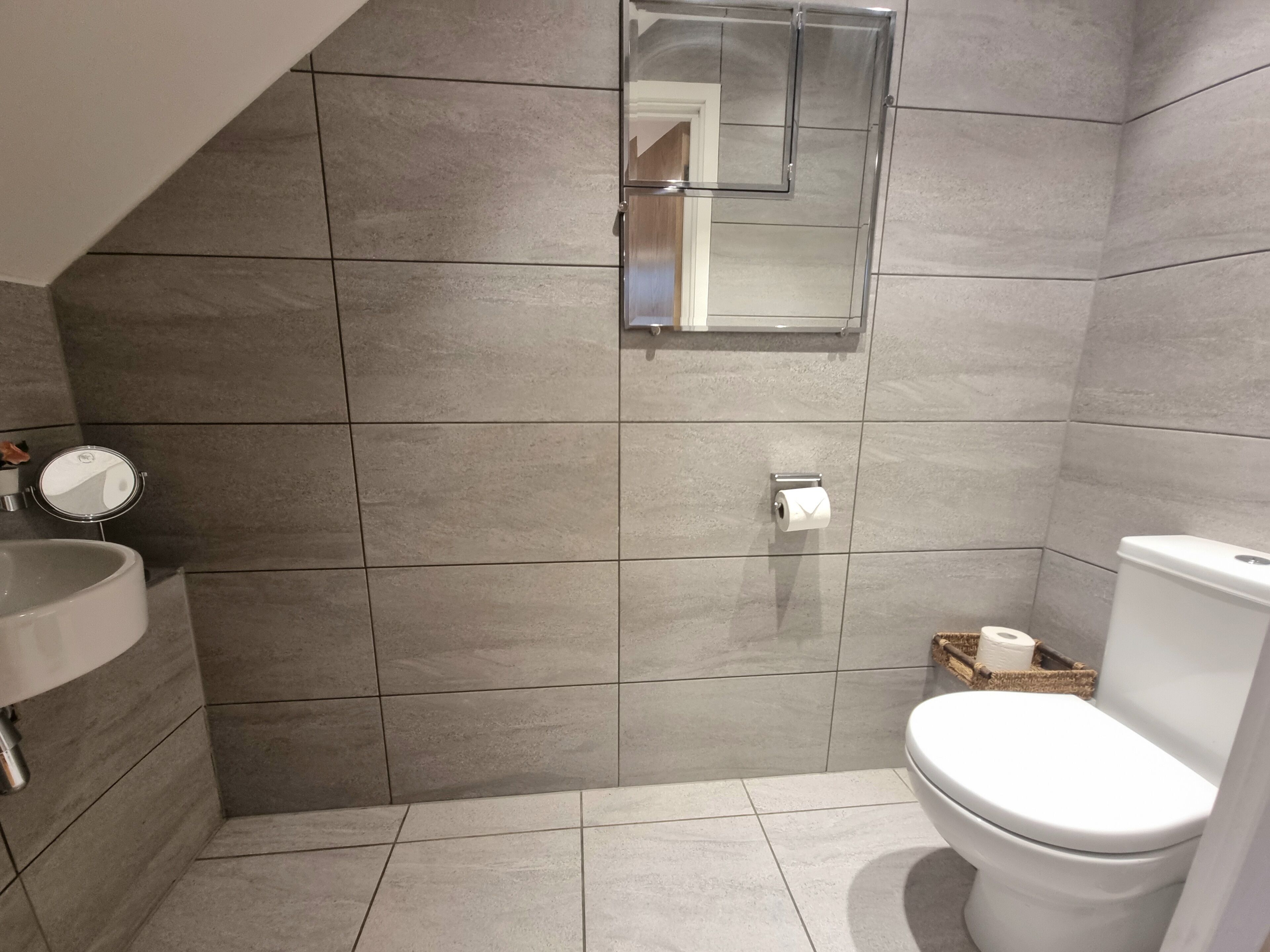Een badkamer met grijze tegels, een wit toilet, een wastafel en een spiegel.