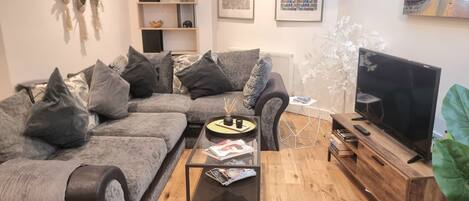 A modern living room with a grey sectional sofa, a glass coffee table, a wooden TV stand, and a large potted plant.