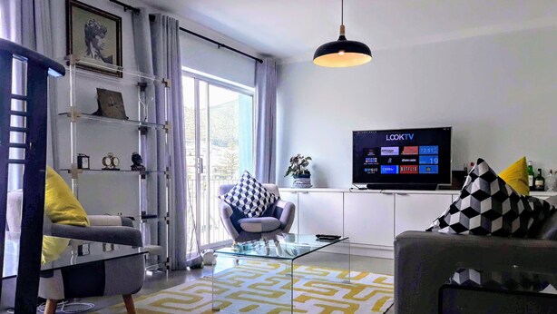 A modern living room with a sofa, armchair, and a glass coffee table. There is a television mounted on the wall and a window with curtains. - Sea Point; Secure 100m² top floor 2 bed apartment with mountain views (Sea Point)