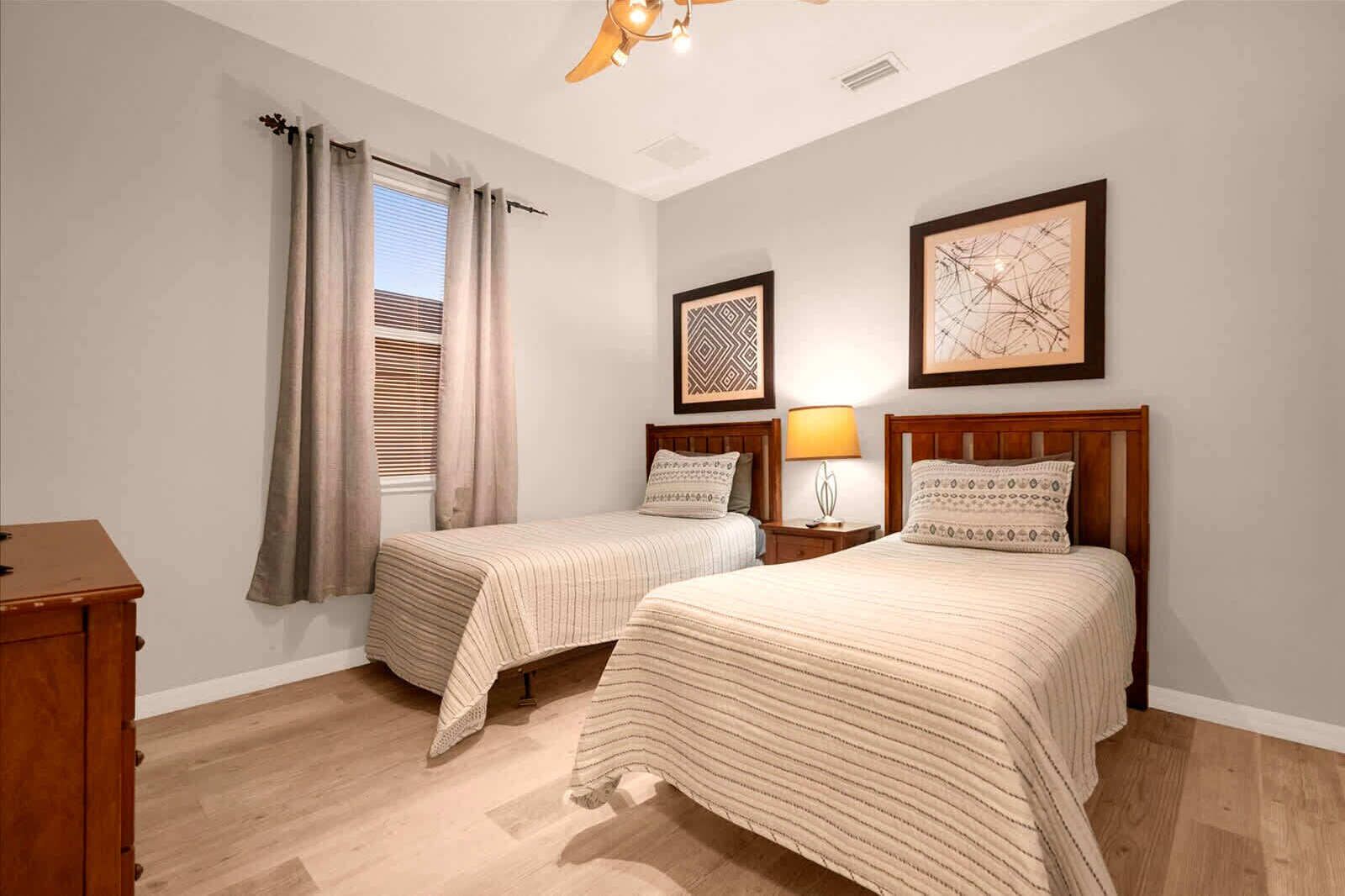 Une chambre avec deux lits simples, une commode en bois, une table de chevet avec une lampe et deux œuvres d’art encadrées accrochées au mur.