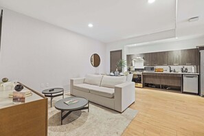 Una sala de estar moderna con un sofá, una mesa de centro y una mesita auxiliar. Se ve una cocina al fondo, con estufa, lavaplatos y armarios.
