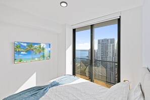 A bedroom with a bed, a painting of a beach, and a view of a cityscape through a large window.