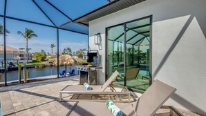 A modern outdoor patio with a grill, lounge chairs, and a glass wall overlooking a body of water. - Villa Serenity - newly built on water, pool & spa (Cape Coral)