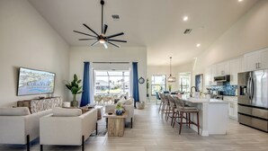 A spacious living room with a sofa set, a coffee table, and a TV on the wall. There is a kitchen area with a refrigerator, microwave, and bar stools. - Villa Serenity - newly built on water, pool & spa (Cape Coral)