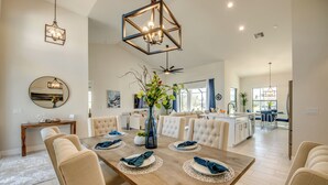 A spacious dining area with a wooden table set for six, surrounded by beige upholstered chairs. A large mirror and pendant lights are visible on the walls. - Villa Serenity - newly built on water, pool & spa (Cape Coral)