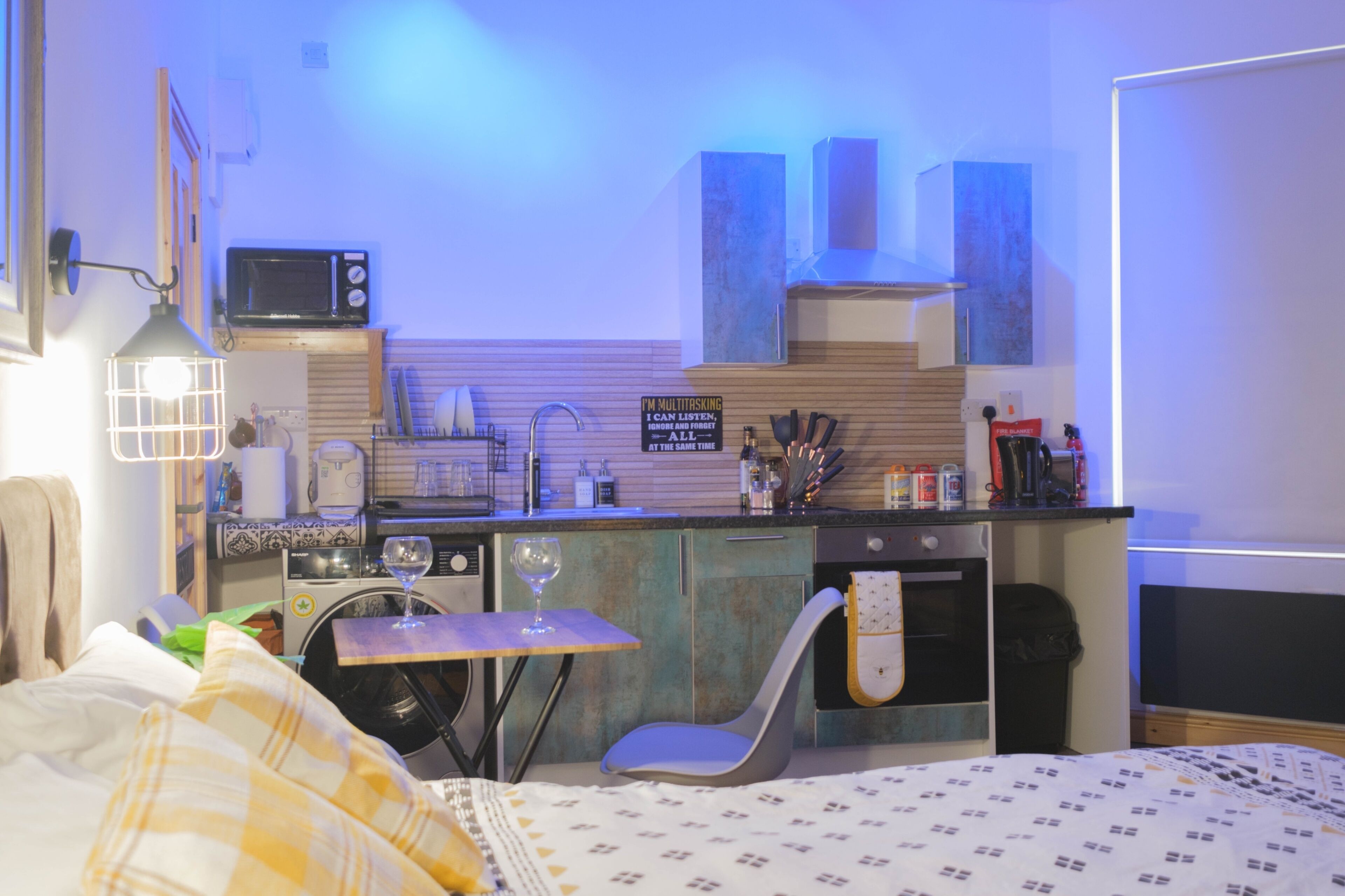 A compact kitchen with a microwave, sink, and a sign that says "I'm multitasking." There's a small table with a glass, a chair, and a bed with a checkered blanket in the foreground.