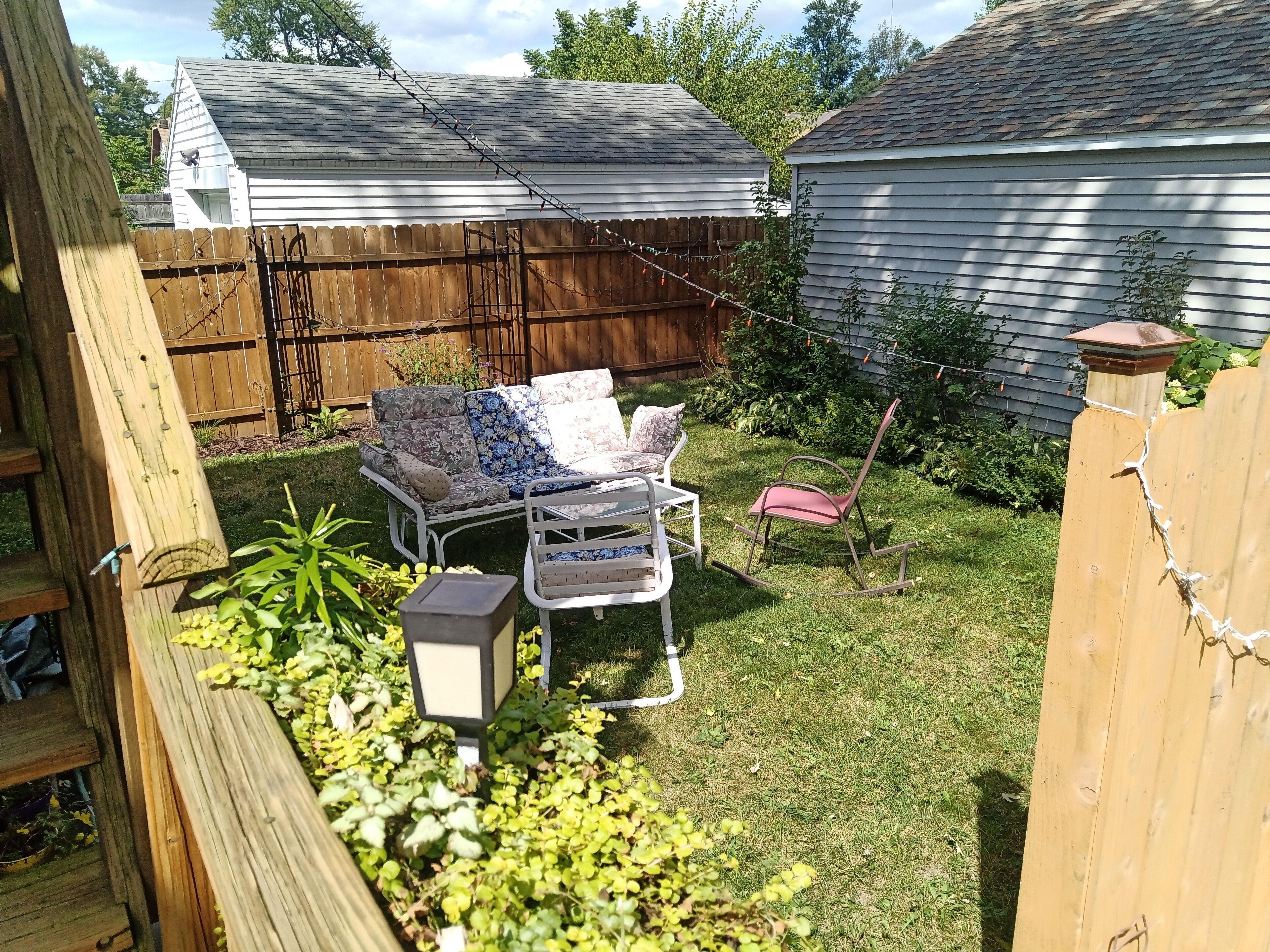 Un jardin avec une terrasse en bois, une pelouse, une chaise en métal blanche, une chaise berçante et un petit espace aménagé en jardin avec des plantes.