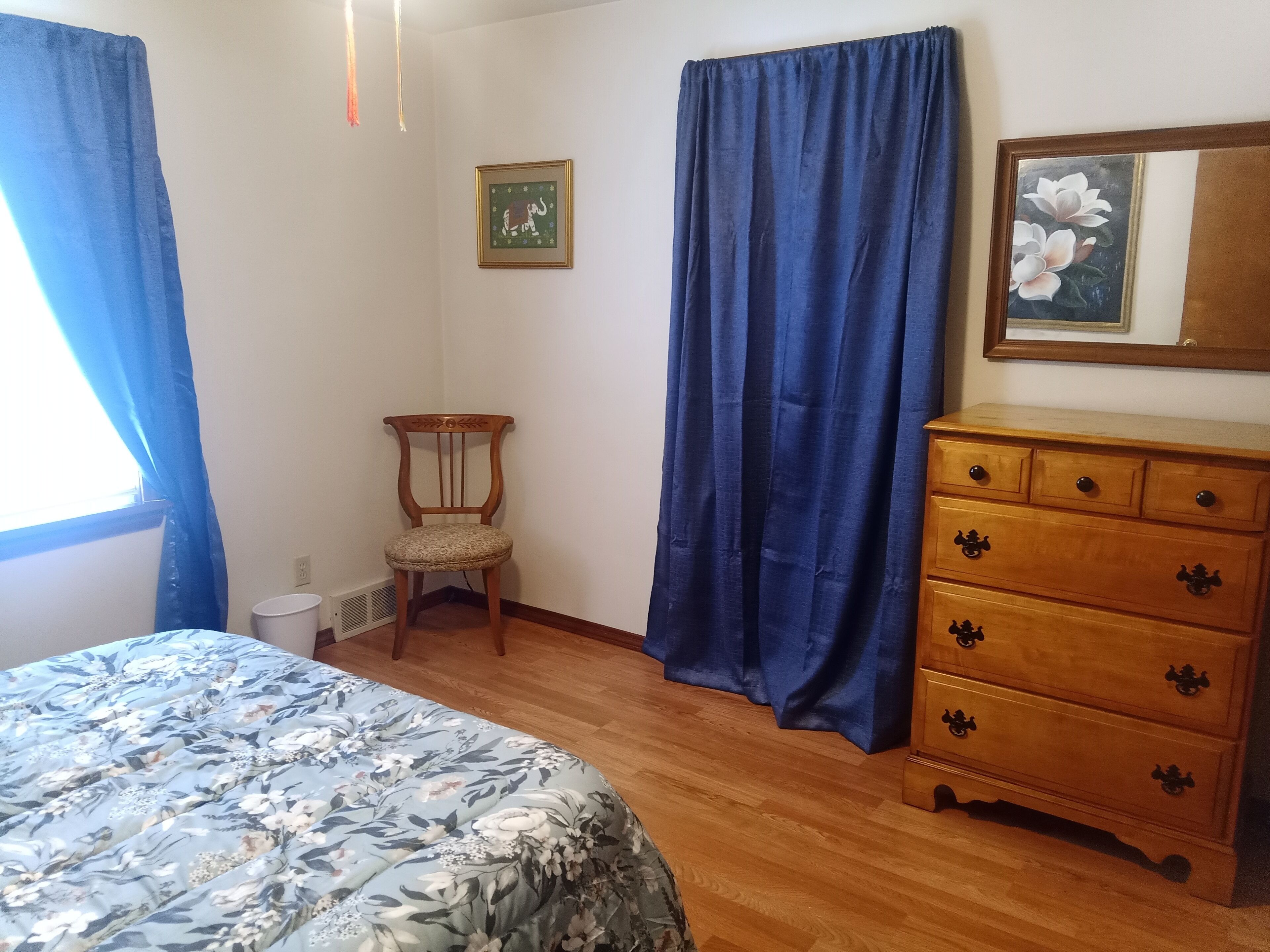 Una camera da letto con una cassettiera in legno, una sedia, un letto con copriletto floreale, tende blu e un quadro appeso al muro.