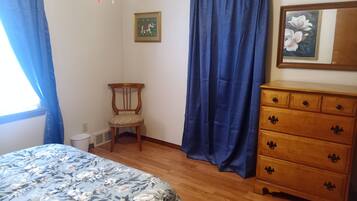 Una camera da letto con una cassettiera in legno, una sedia, un letto con copriletto floreale, tende blu e un quadro appeso al muro.