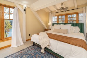 Une chambre avec un grand lit, une tête de lit verte, un ventilateur de plafond et une vue sur les arbres par les fenêtres.