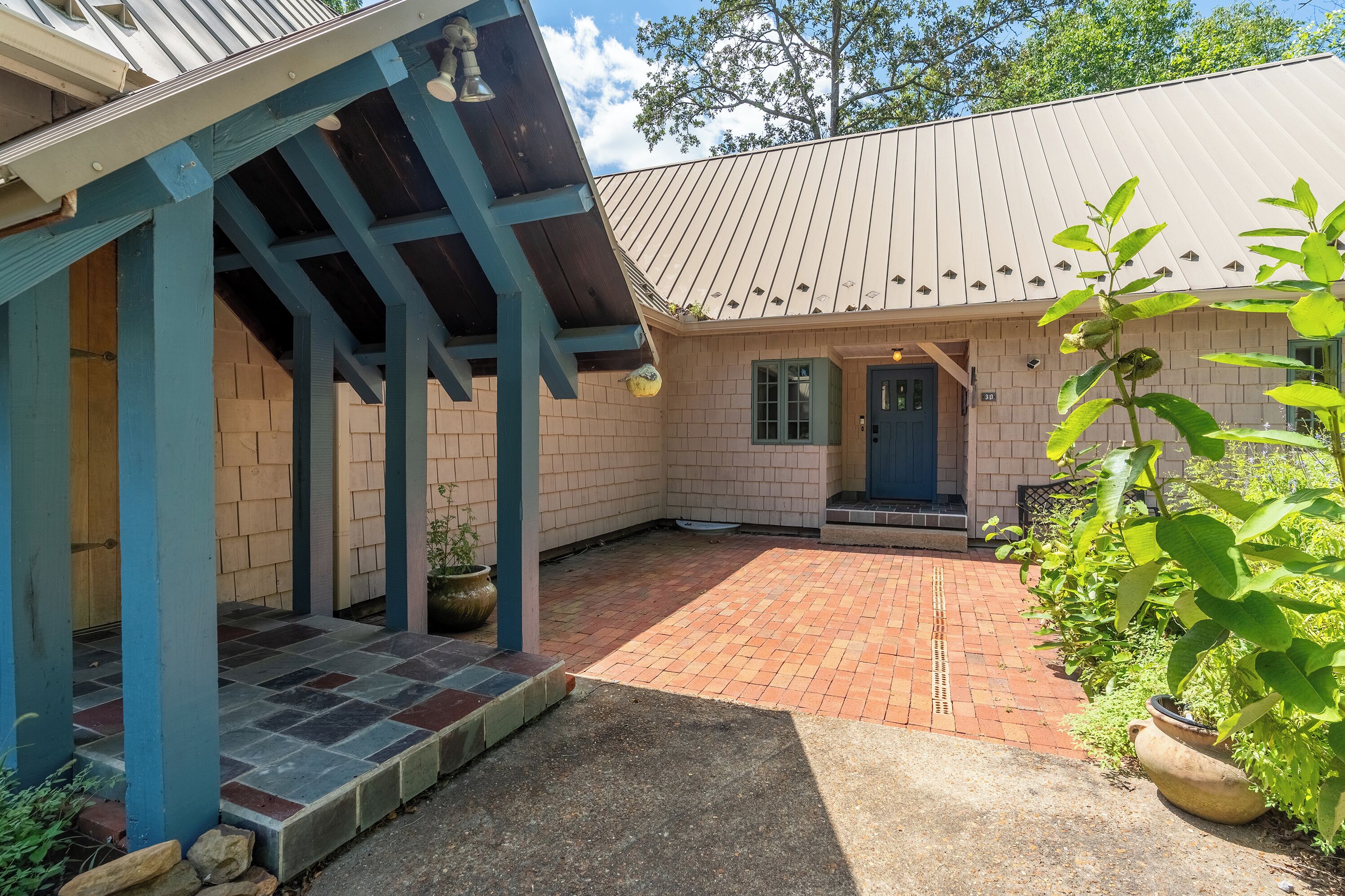 Un patio couvert doté d’un sol en briques, d’une structure en bois bleue et d’une porte donnant accès à un bâtiment dont le toit est recouvert de tuiles.