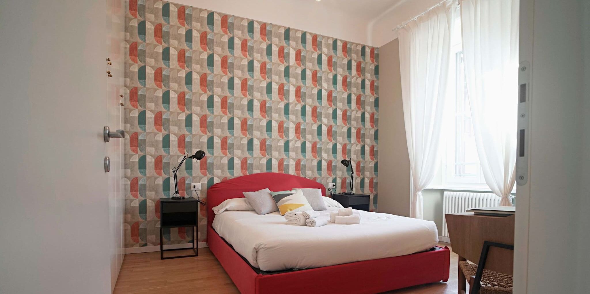 Ein Schlafzimmer mit einem roten Bett, einem Wandteppich mit Muster, einem Nachttisch mit Lampe und einem Fenster mit Vorhängen.