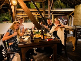 Um grupo de pessoas sentadas à mesa com comida e bebidas, um cão, e uma placa com a inscrição 'Co Palm & Lounge'.