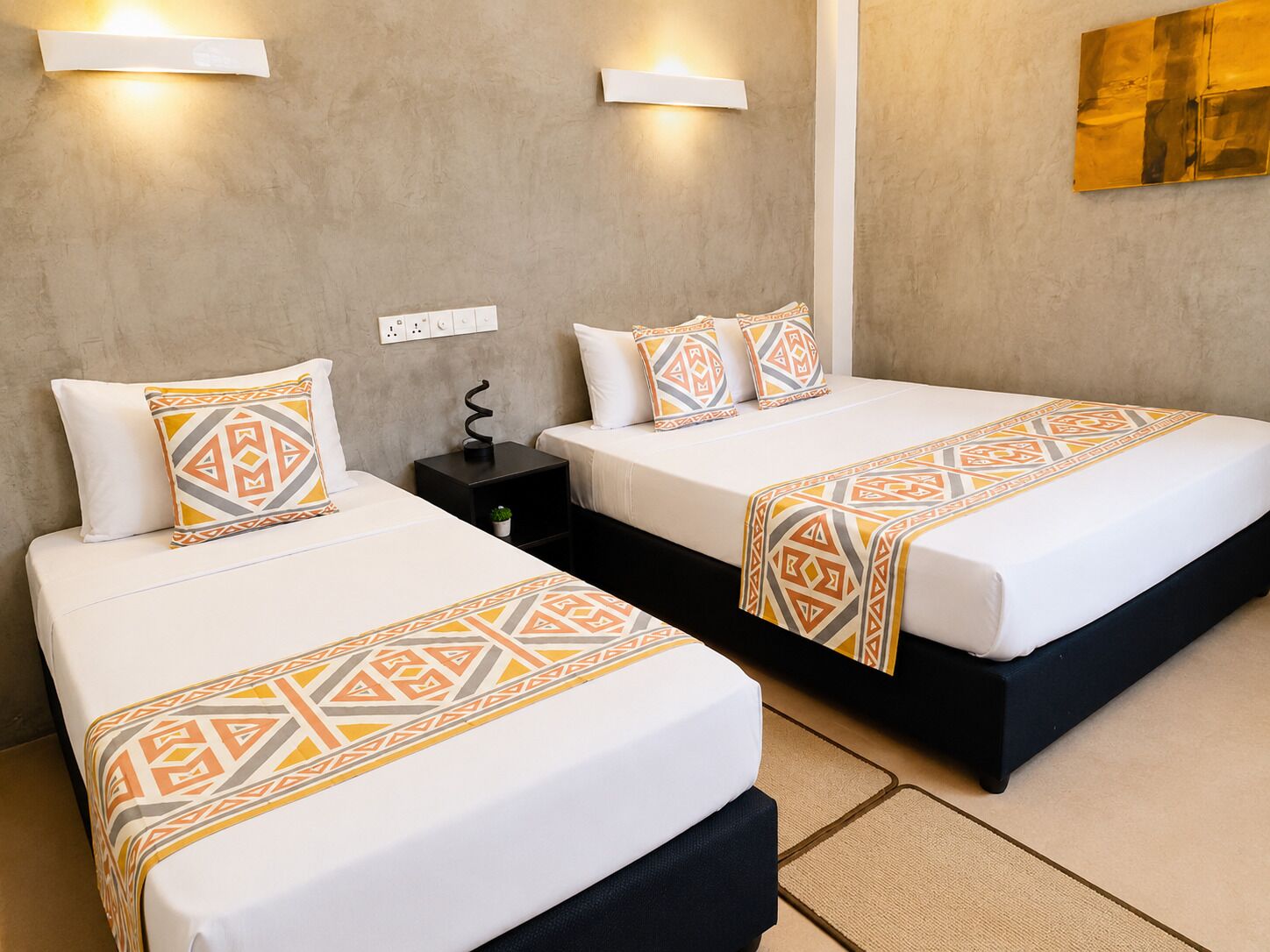 A hotel room with two beds, each with a patterned bedspread and matching pillows. There are wall-mounted lights and a small black nightstand with a plant.