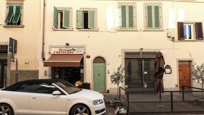 Une voiture décapotable blanche est garée sur une rue pavée, devant un immeuble dont la porte est peinte en vert et sur lequel on peut lire l’inscription « L’Ortolano ».