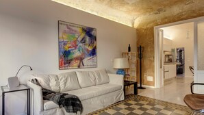 Un salon avec un canapé blanc, un tapis à motifs, un tableau accroché au mur et une cuisine en arrière-plan.