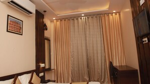 A hotel room with a bed, a desk, a ceiling fan, an air conditioning unit, a television, and curtains. - Hotel Aishwarya Inn (Jaipur)