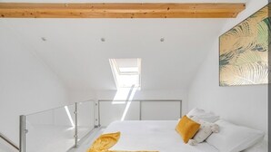 Une chambre avec un plafond en pente, un puits de lumière, un lit avec une literie blanche et un plaid jaune, et un tableau accroché au mur.