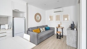Un espace de vie compact comprenant un coin cuisine, un canapé gris agrémenté de coussins colorés, une petite table en bois et un téléviseur posé sur un meuble.