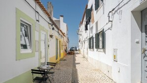 Une ruelle étroite pavée de pierres, avec des bâtiments de chaque côté, une table entourée de chaises devant un bâtiment blanc, et une voiture garée au loin.