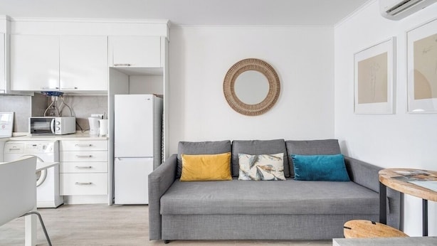 Une cuisine moderne avec des meubles blancs, un canapé gris agrémenté de coussins colorés et un miroir rond accroché au mur.