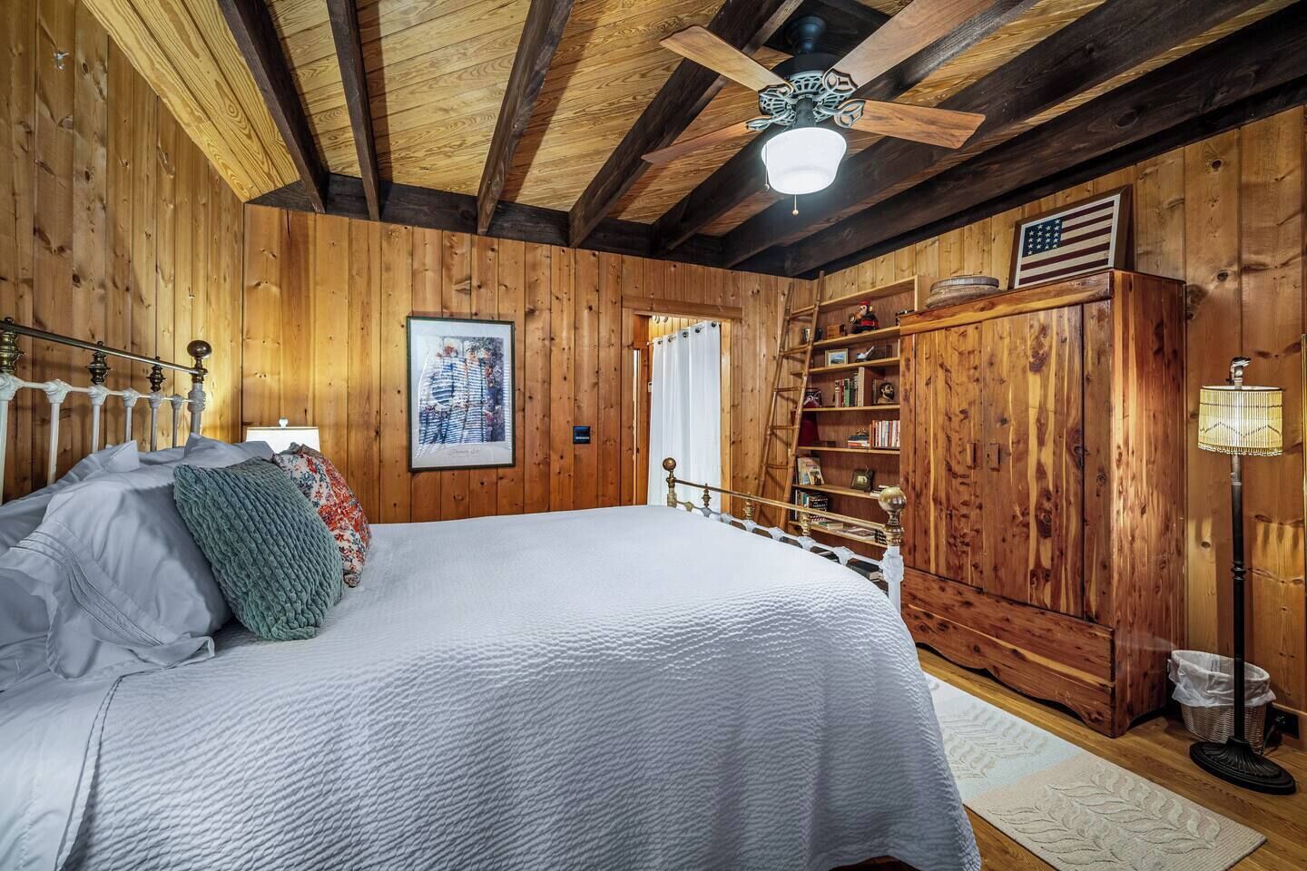 A wooden cabin bedroom with a bed, a ceiling fan, a bookshelf, and a framed picture on the wall.