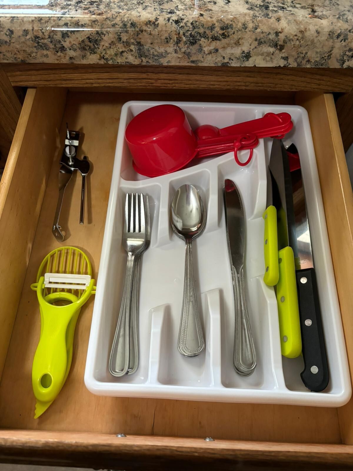 A drawer with a utensil organizer containing a red measuring cup, a red spatula, a yellow grater, a knife, a spoon, a fork, and a pair of tongs.