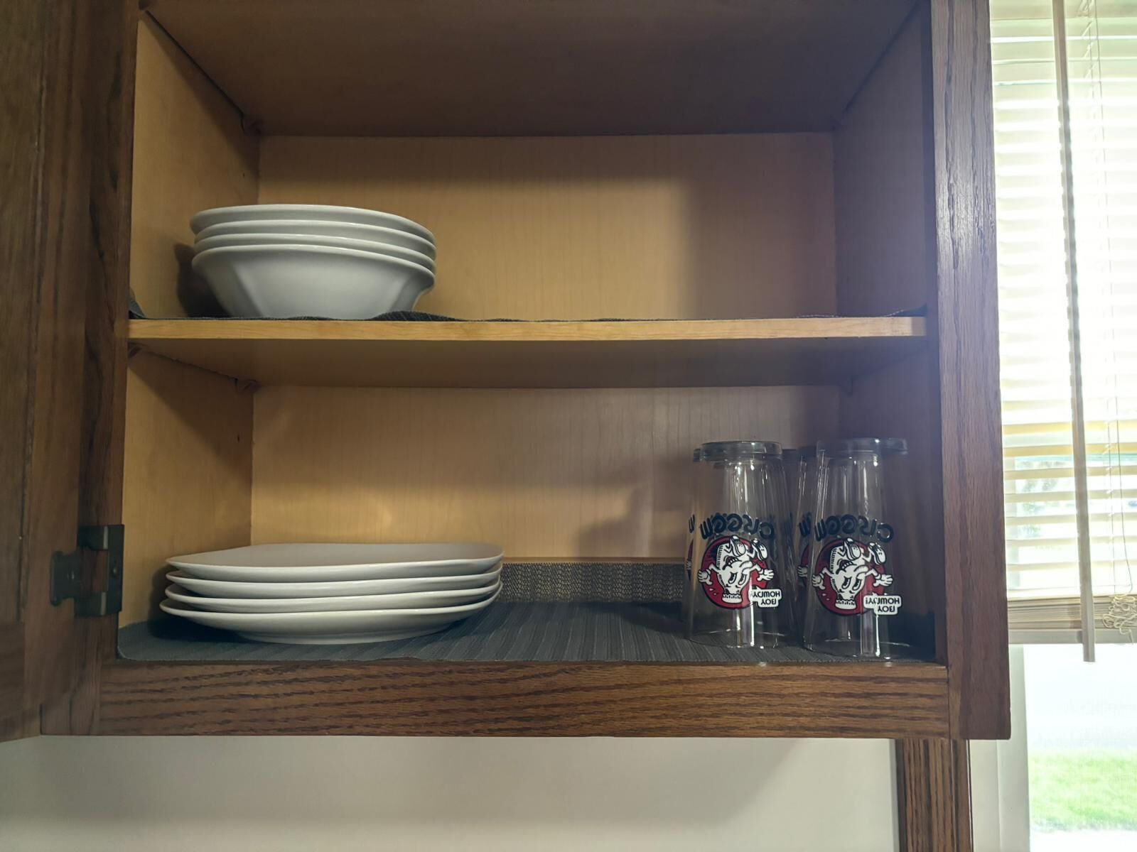 A wooden cabinet with two shelves; the top shelf holds white plates and the bottom shelf has clear glass bottles with labels.