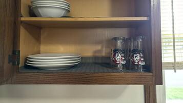 A wooden cabinet with two shelves; the top shelf holds white plates and the bottom shelf has clear glass bottles with labels.