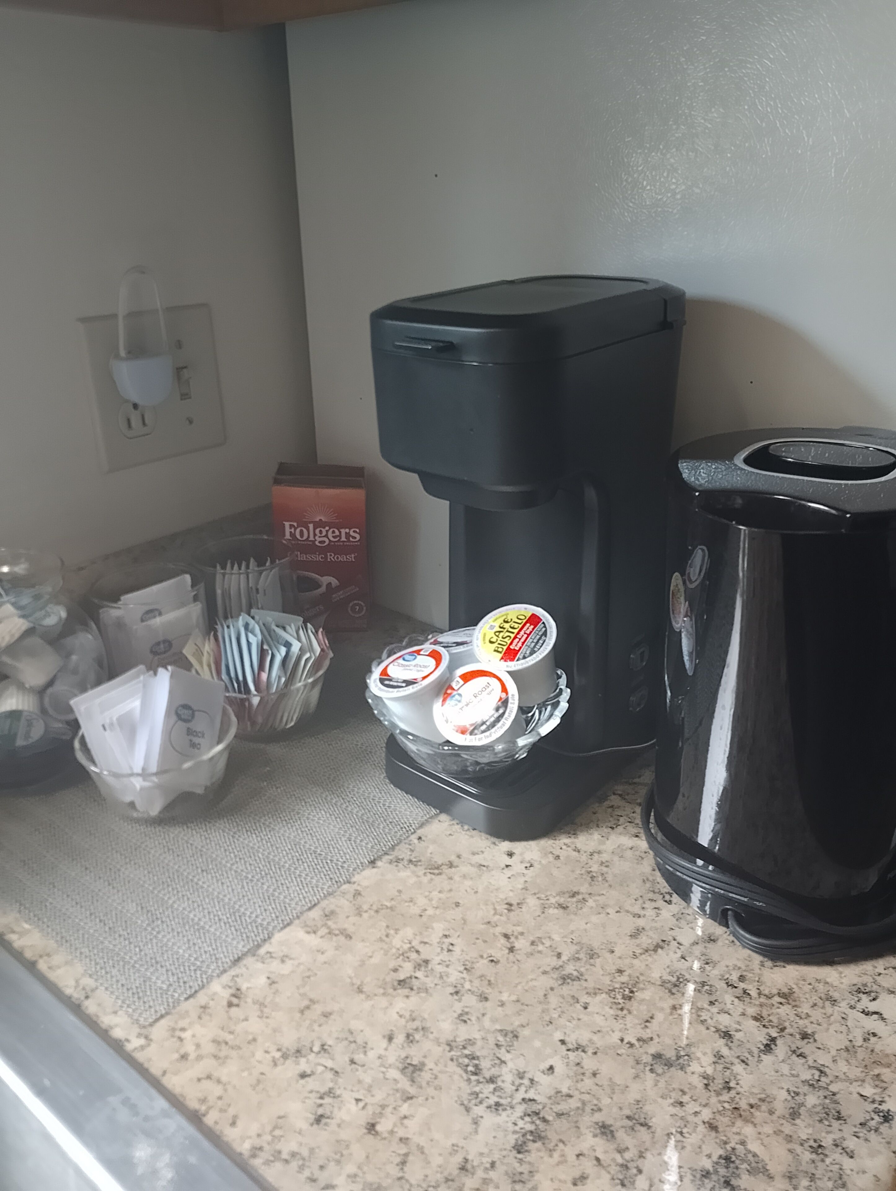 Eine Kaffeemaschine mit Kaffee-Kapseln, ein schwarzer Wasserkocher und eine Packung Folgers Classic Roast Kaffee.