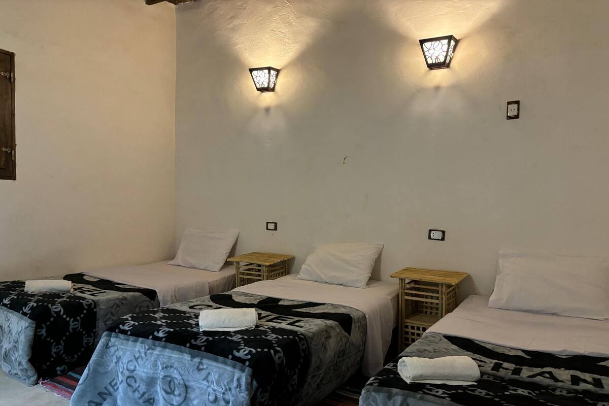 Ein Zimmer mit drei Einzelbetten, jedes mit einem weißen Kissen und einer geblümten Decke. An der Wand über den Betten befinden sich zwei Wandleuchten.