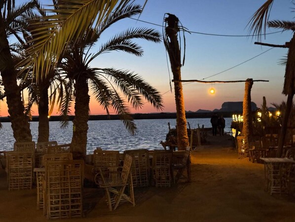 Een zithoek aan het strand met rieten meubilair en palmbomen, met uitzicht op een kalme zee bij zonsondergang.