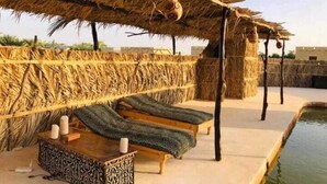 A thatched-roof outdoor seating area with two lounge chairs and a table, overlooking a pool. - Gazrashek Siwa Eco Lodge (Siwa)