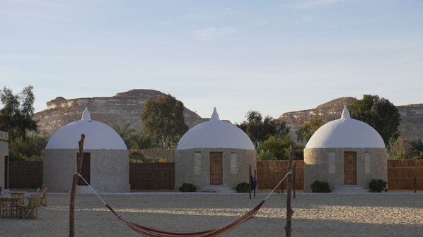 Three dome-shaped buildings with doors, set against a backdrop of rocky cliffs and trees. - Gazrashek Siwa Eco Lodge (Siwa)