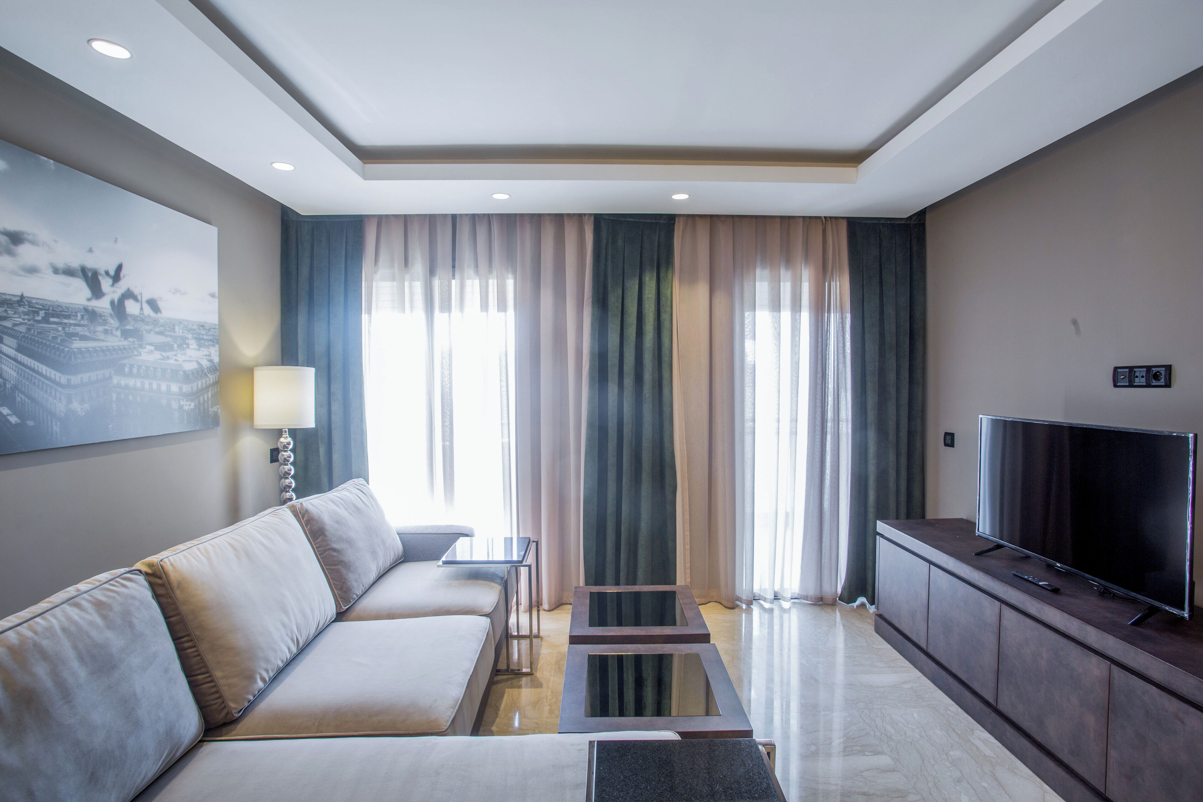 A modern hotel room with a sofa, a flat-screen TV, a small table, and a large window with curtains.