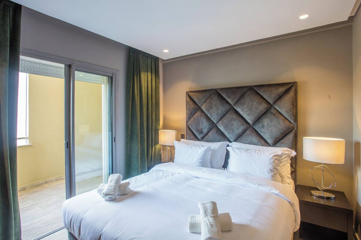Ein Hotelzimmer mit einem großen Bett, zwei Nachttischlampen, einem modernen Kopfteil und Blick durch eine Schiebetür.