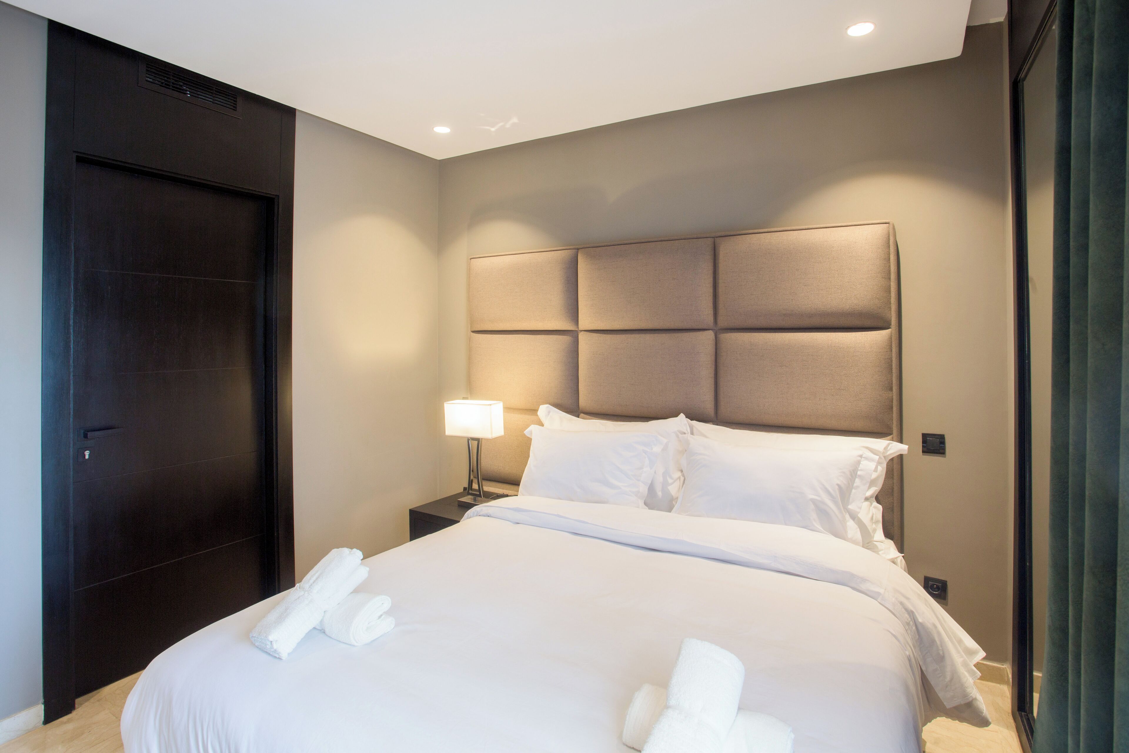 A hotel room with a large bed, a dark wooden door, a nightstand with a lamp, and a headboard with a light panel.