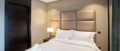 A hotel room with a large bed, a dark wooden door, a nightstand with a lamp, and a headboard with a light panel.