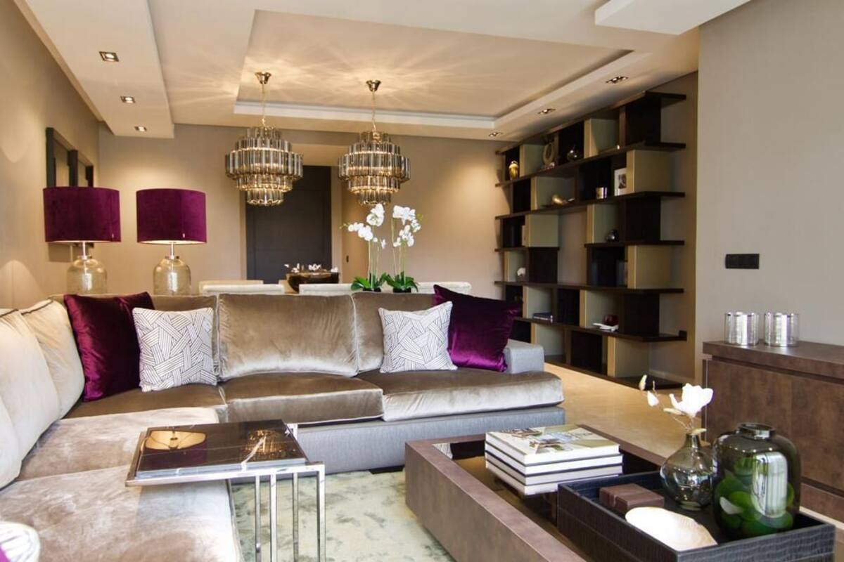 Ein modernes Wohnzimmer mit einem grauen Ecksofa, zwei violetten Lampen, einer Kristallleuchter und einem Bücherregal.