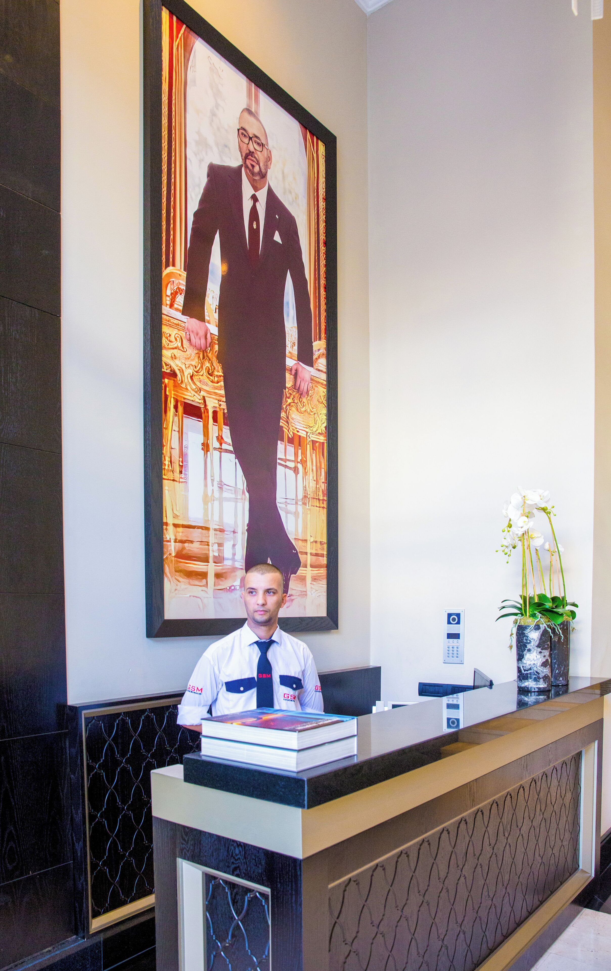 A hotel reception area with a large framed portrait of a man in a suit, a reception desk, and a vase with white flowers.