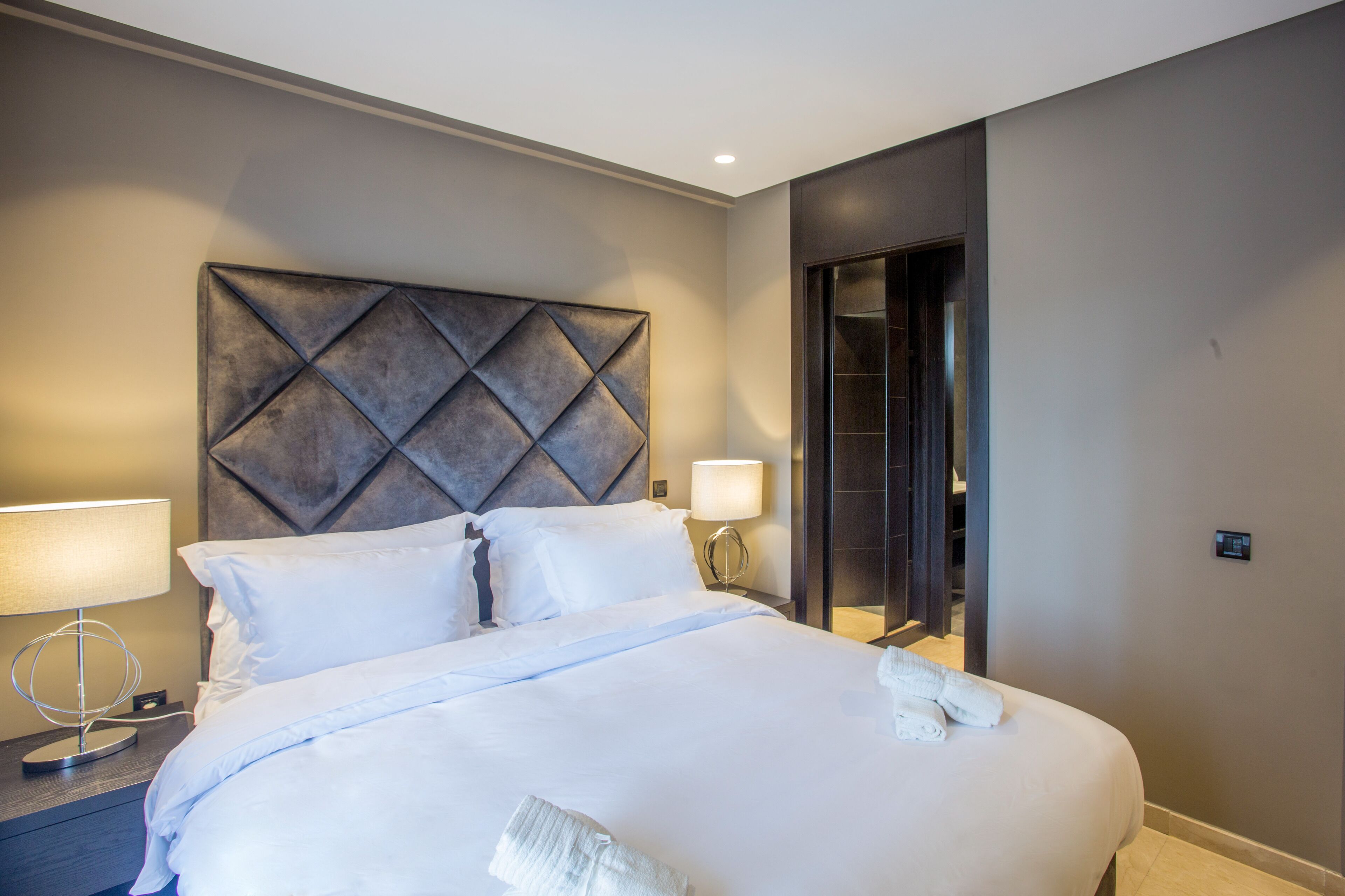 A modern hotel room with a large bed, a headboard with a geometric pattern, bedside lamps, and a walk-in closet.