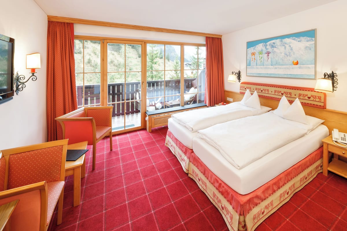A hotel room with two beds, a desk, a chair, a TV, and a balcony with a view of trees and mountains.