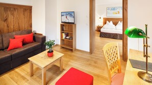 Une chambre d’hôtel moderne avec une tête de lit en bois, un canapé agrémenté de coussins rouges, une petite table en bois sur laquelle est posée une plante en pot, une bibliothèque et un bureau avec une lampe de bureau verte.