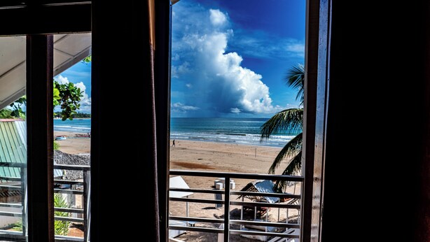 A beach view from a window with a metal railing, palm trees, and a clear sky.