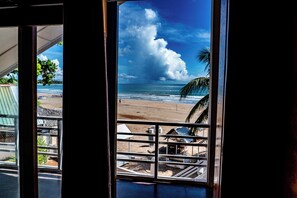 Une vue sur la plage, prise depuis une fenêtre ornée d’une rambarde en métal, avec des palmiers et un ciel dégagé.
