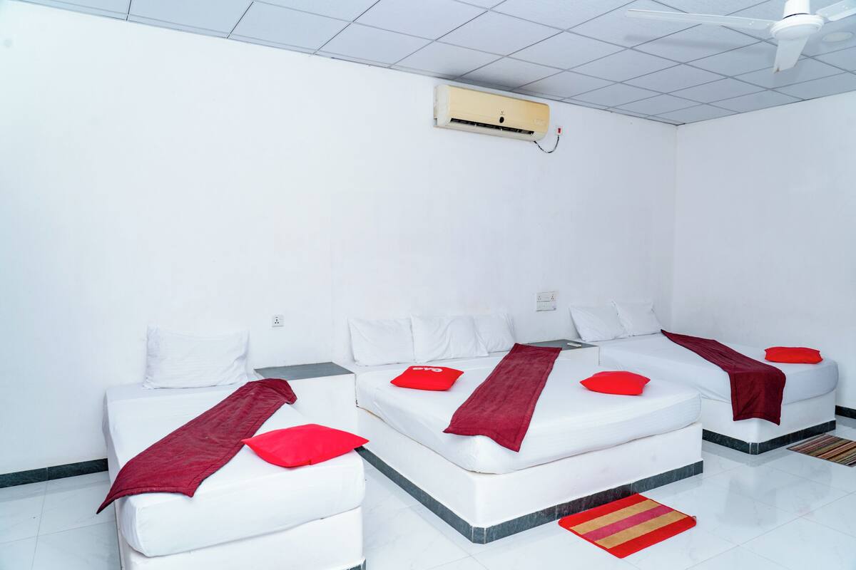 A hotel room with two beds, white bedding, red pillows, a red throw blanket, a ceiling fan, and an air conditioning unit.