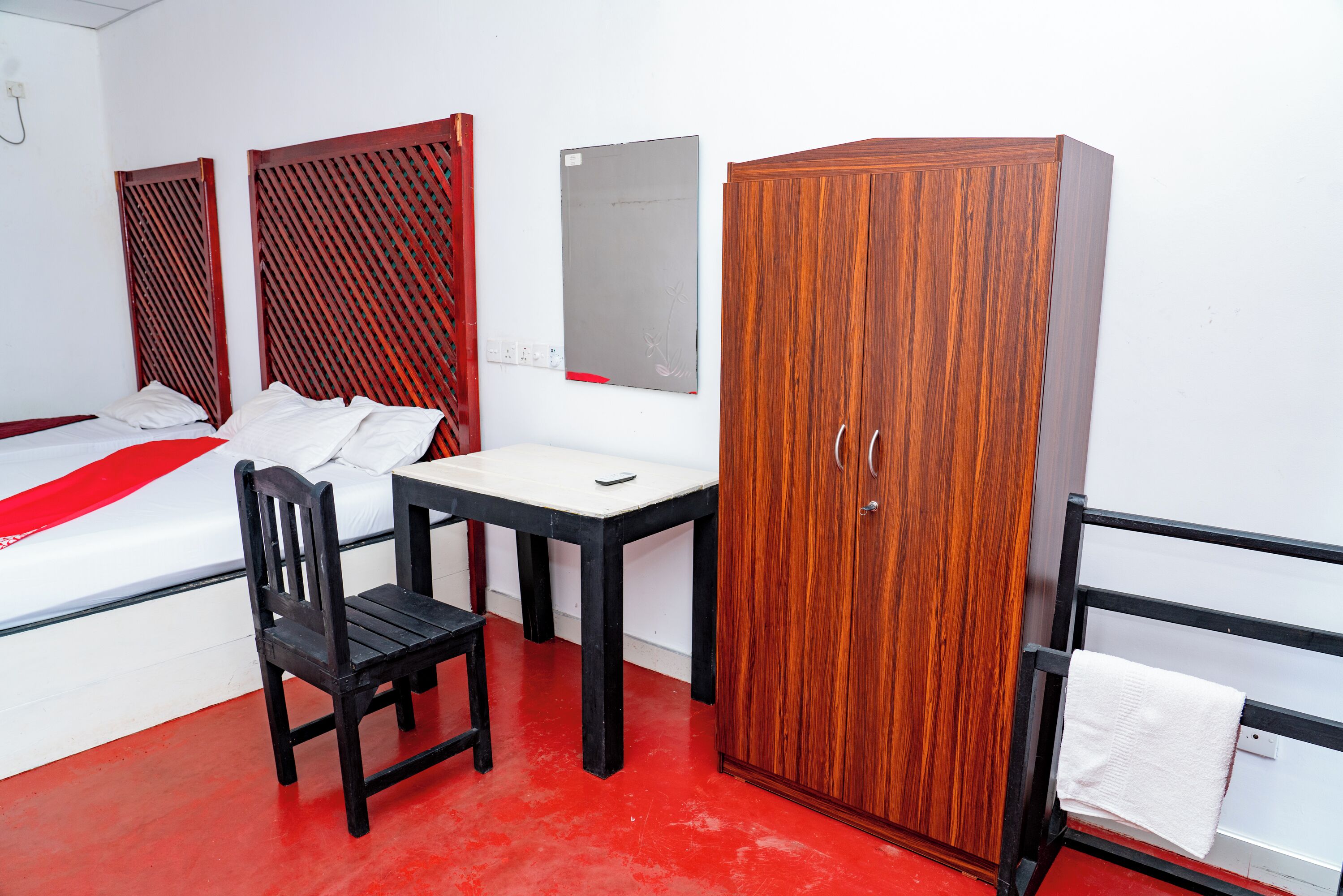 A hotel room with a bed, a wooden wardrobe, a small table with a chair, and a red floor.