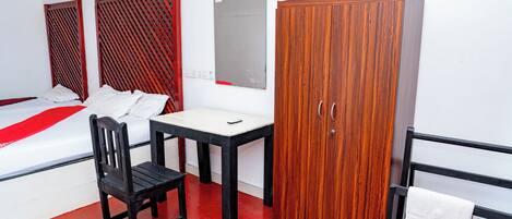 A hotel room with a bed, a wooden wardrobe, a small table with a chair, and a red floor.